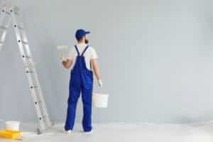 Professional painter in blue overalls painting an interior wall with a roller.
